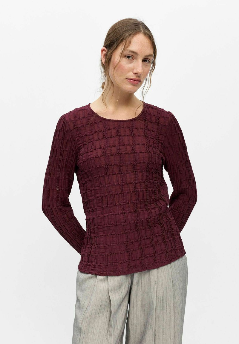 Femme aux cheveux châtain clair portant un haut bordeaux texturé à manches longues et un pantalon large gris clair, posant devant un fond blanc.