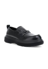 Mocasín de cuero negro con suela gruesa y estriada, detalle decorativo de metal en el empeine y tirador en el talón, mostrado sobre fondo blanco.