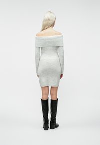 Kvinne med langt blondt hår, iført en grå off-shoulder strikket kjole og svarte knelange høye hæler, står vendt bort fra kameraet mot en hvit bakgrunn.