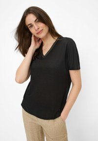 Sort kortærmet V-hals top i let materiale, med en subtil glans og kontrasterende stribet halsudskæring; båret med beige linnedbukser.