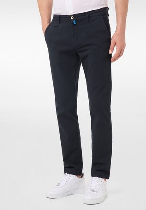 Mann trägt dunkelblaue, schmal geschnittene Hose und weiße Sneakers, steht mit den Händen in den Taschen vor einem weißen einfarbigen Hintergrund.