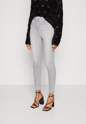 Persoon die lichtgrijze skinny jeans, een zwarte, pluizige trui met lange mouwen en zwarte sandalen met bandjes en blokhak draagt, staand tegen een witte achtergrond.
