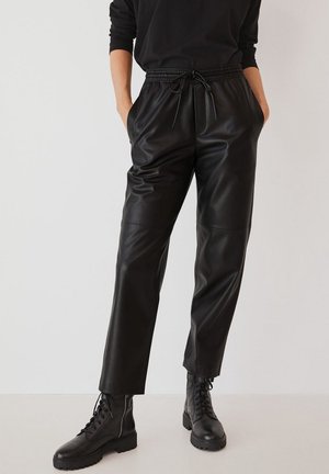 Osoba nosząca czarne joggery ze sztucznej skóry z ściągaczem w talii i czarne wojskowe buty sznurowane, stojąca na jednolitym tle.