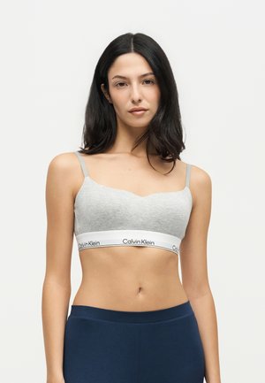 Жена с дълга тъмна коса, носеща светлосив спортен сутиен Calvin Klein и тъмносини панталони, гледаща напред на едноцветен фон.