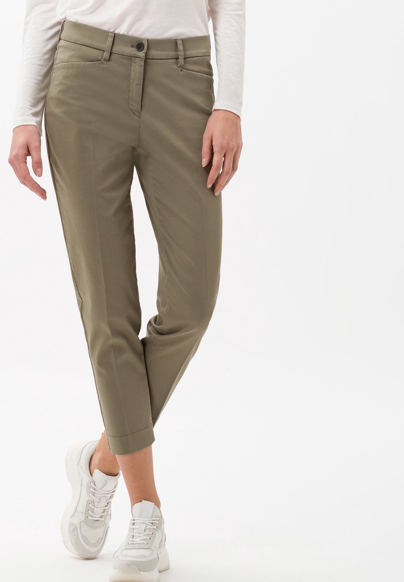Femme portant un pantalon court vert olive et des baskets blanches debout devant un fond blanc uni.