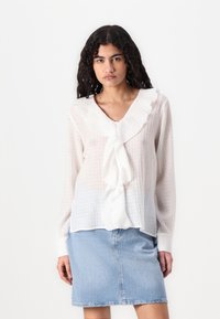 Blusa blanca de manga larga con escote en V con volantes y tejido texturizado, combinada con una falda de mezclilla azul clara.