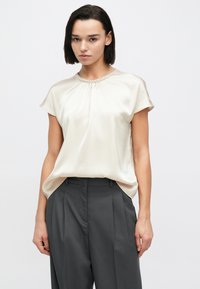 Επάνω μέρος από σατέν σε ανοιχτό μπεζ χρώμα με κοντά μανίκια, σούρες στο ντεκολτέ και περίτεχνη διακόσμηση με χάντρες. Συνδυάζεται με σκούρο γκρι ψηλόμεσα παντελόνια.