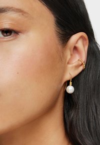 Primo piano dell'orecchio di una donna con capelli lunghi e scuri, che indossa un orecchino a manetta dorato con perline e un orecchino a gancio dorato con una grande perla bianca.