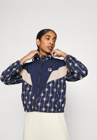 Giacca blu navy con accenti beige, caratterizzata da un motivo geometrico. Colletto alto e polsini elasticizzati, chiusura con zip e dettaglio del logo sul petto.