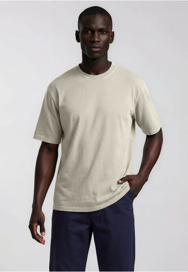 Homme à la peau foncée portant un t-shirt beige ample à manches courtes et un pantalon bleu marine foncé, debout devant un fond clair uni.
