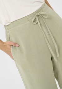 Svetlozelené, voľné nohavice s viazaním v páse a bočnými vreckami. Hladká textúra so subtílnymi stehovými detailmi.