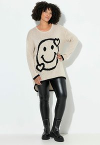 Beige gebreide trui met een grote zwarte glimlachende gezichtafdruk, gedragen met zwarte glanzende leggings en gestifte zwarte laarzen. Lange, relaxte pasvorm.