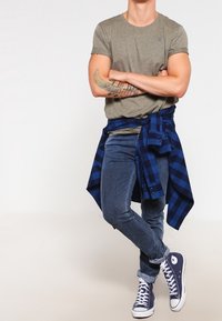 Camiseta verde oliva, camisa de cuadros azul anudada a la cintura, vaqueros azul oscuro y zapatillas Converse azul marino con suelas blancas.