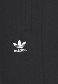Černé bavlněné joggery s bílým logem Adidas a designem tří listů, s horizontálními proužky nad názvem značky. Jemná textura.