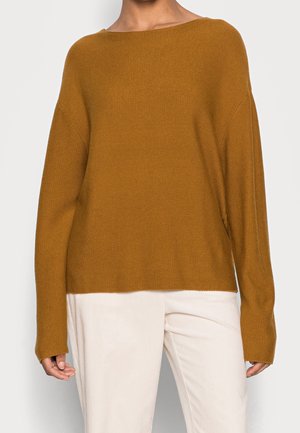 Osoba ubrana w luźny, długi sweter z dzianiny w kolorze musztardowym z dekoltem w łódkę, w połączeniu z jasnobeżowymi spodniami.