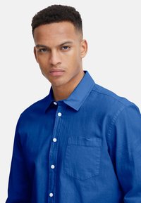 Hemd in Hellblau aus Baumwolle mit einer Tasche auf der linken Seite, ausgestattet mit einem Kragen und weißen Knöpfen. Glatte Textur, kurze Ärmel.