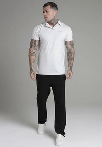 Vit pikétröja med krage och diskret logotyp, kombinerad med svarta byxor och vita sneakers. Modellen har tatueringar och neutral bakgrund.