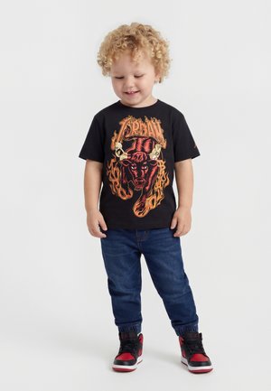 Uśmiechnięte małe dziecko z kręconymi blond włosami, ubrane w czarny T-shirt z grafiką płonącego byka, niebieskie dżinsy oraz czarno-czerwone trampki, stojące na białym tle.