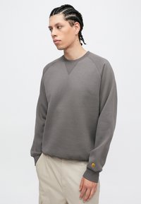 Gray sweatshirt with a round neck and raglan sleeves. Ribbed cuffs and hem, featuring a small orange logo on the left sleeve.