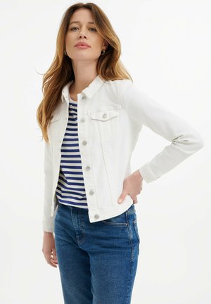 Giacca di jeans bianca con chiusura a bottoni, due tasche sul petto e maniche lunghe, indossata sopra una camicia a righe blu e bianche e jeans blu.