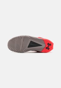 Egy sportcipő talpa szürke mintázattal és piros sarkával, fekete Under Armour logóval, fehér háttéren bemutatva.
