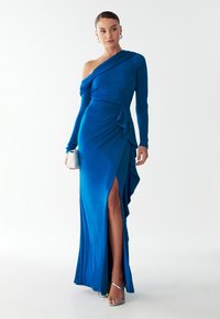 Ühe õlaga, slim-fit sinine kleit pehmest kangast, millel on volangide detail ja reie kõrgune lõhik, kombineeritud hõbedaste rihmaga kontsakingadega.