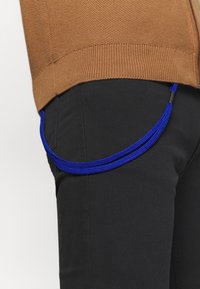 Vue de côté d'une personne portant un pull en maille marron et un pantalon noir avec un cordon tressé bleu vif qui pend de la poche.