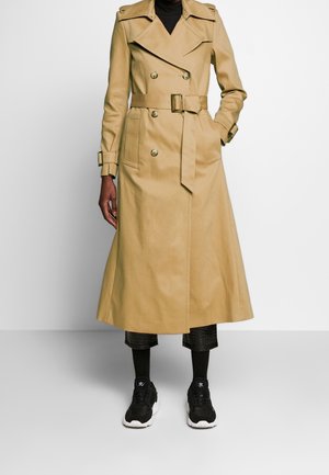 Personne portant un long trench-coat beige avec ceinture, un pantalon noir raccourci, des chaussettes noires et des baskets noires, debout devant un fond uni.