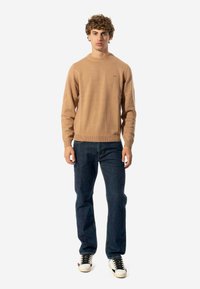 Maglione a maglia beige con collo rotondo, abbinato a jeans in denim blu scuro. Il maglione presenta un logo discreto sul petto e orlo a costine.