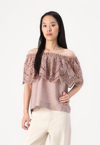 Top off-the-shoulder in mauve chiaro, con un sovrapposto di pizzo scallopato con tagli intricati, abbinato a pantaloni larghi color crema.