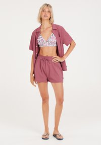 Dvoudílný plavecký outfit s vzorovanou podprsenkou a texturovanými, volnými růžovými šortkami. Modelka nosí žabky. Jednoduchý, uvolněný design.