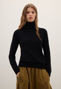 Schwarzer Rollkragenpullover aus weichem Material, mit langen Ärmeln und figurbetontem Schnitt, kombiniert mit einem strukturierten, senffarbenen Rock.