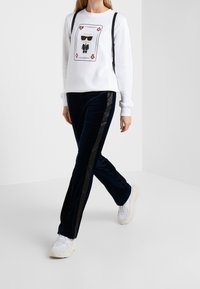 Sudadera blanca con una impresión gráfica que presenta a un personaje con gafas de sol, combinada con pantalones de terciopelo azul marino con franjas laterales negras. Zapatillas blancas.