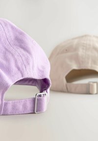 Két állítható baseball sapka: az egyik levendula színű, a másik bézs. Mindkettő puha szövet felülettel rendelkezik, és fém alkatrész a méretállításhoz hátul.