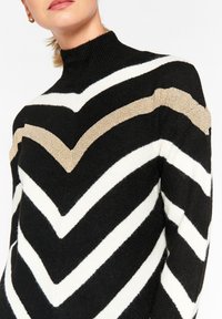 Suéter de punto en blanco y negro con patrón de chevron, cuello alto y una franja dorada como acento. Textura suave y diseño ajustado.