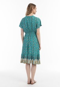 Rochie turcoaz cu flori, cu mâneci scurte, talie elastică și tiv cu un model care include accente galbene și albe. Material ușor.