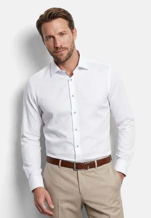 Mann mit kurzen braunen Haaren und Bart, trägt weißes Hemd mit Knöpfen und beige Hose mit braunem Gürtel, steht vor einer weißen Wand.
