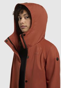 Chaqueta de color óxido con capucha, que cuenta con cierre de cremallera, botones a presión y forro de estampado de leopardo. Fabricada con material liso y resistente al agua.