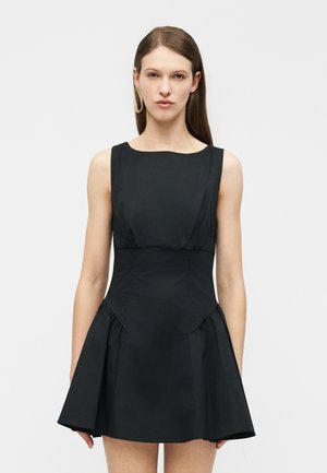 Kvinde iført en sort, ærmeløs kjole med markeret talje og udvidet nederdel, stående foran en ensartet hvid baggrund.