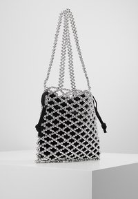 Pochette en tissu noir avec un filet de perles argentées et deux longues anses en perles argentées, exposée sur un piédestal blanc contre un fond uni.