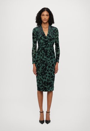 Groene fluwelen jurk met zwart bloemenpatroon, v-hals, lange mouwen, getailleerde silhoutte, kniehoogte, gecombineerd met zwarte stilettohakken.