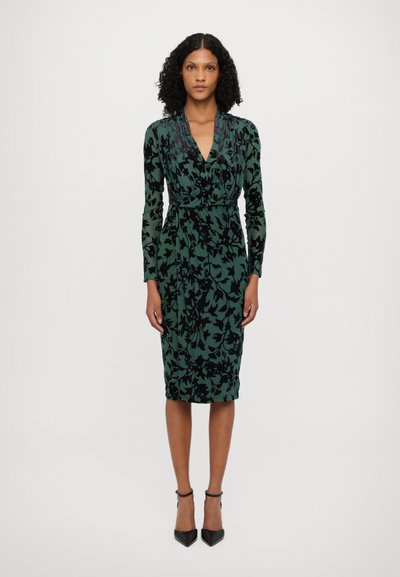 Rochie verde din catifea cu model floral negru, decolteu în V, mâneci lungi, siluetă ajustată, până la genunchi, asortată cu tocuri stiletto negre.