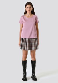 Rosa kurzärmeliges Oberteil kombiniert mit einem grauen karierter, plissierter Rock. Das Model trägt schwarze kniehohe Stiefel. Weiches Material, lockere Passform und dezenter Glanz.