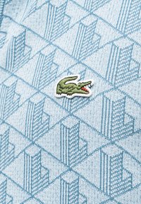 Πράσινο κεντημένο λογότυπο κροκόδειλου σε ανοιχτό μπλε ύφασμα με γεωμετρικό τριγωνικό μοτίβο.