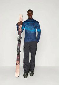 Suusariietus, mis sisaldab sinist, pikkade varrukatega, lukuga ülemist osa lainemustriga, musti pükse ja suuska, mille kinnitused on püstiselt hoitud.