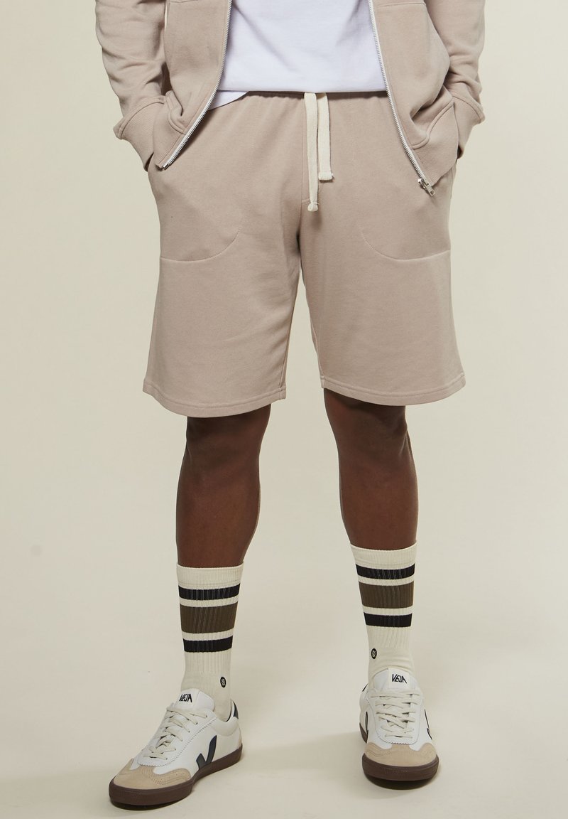 Mann in beigen Kordelzugshorts, beiger Zip-Jacke mit Händen in den Taschen, gestreiften Socken und weißen Veja-Sneakern vor leichtem Hintergrund.
