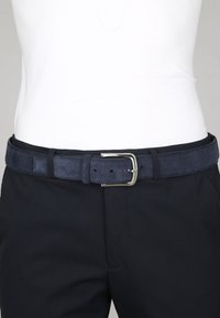 Navyblauwe suède riem met een zilveren rechthoekige gesp, gedragen over zwarte broek en een ingestopt wit shirt.