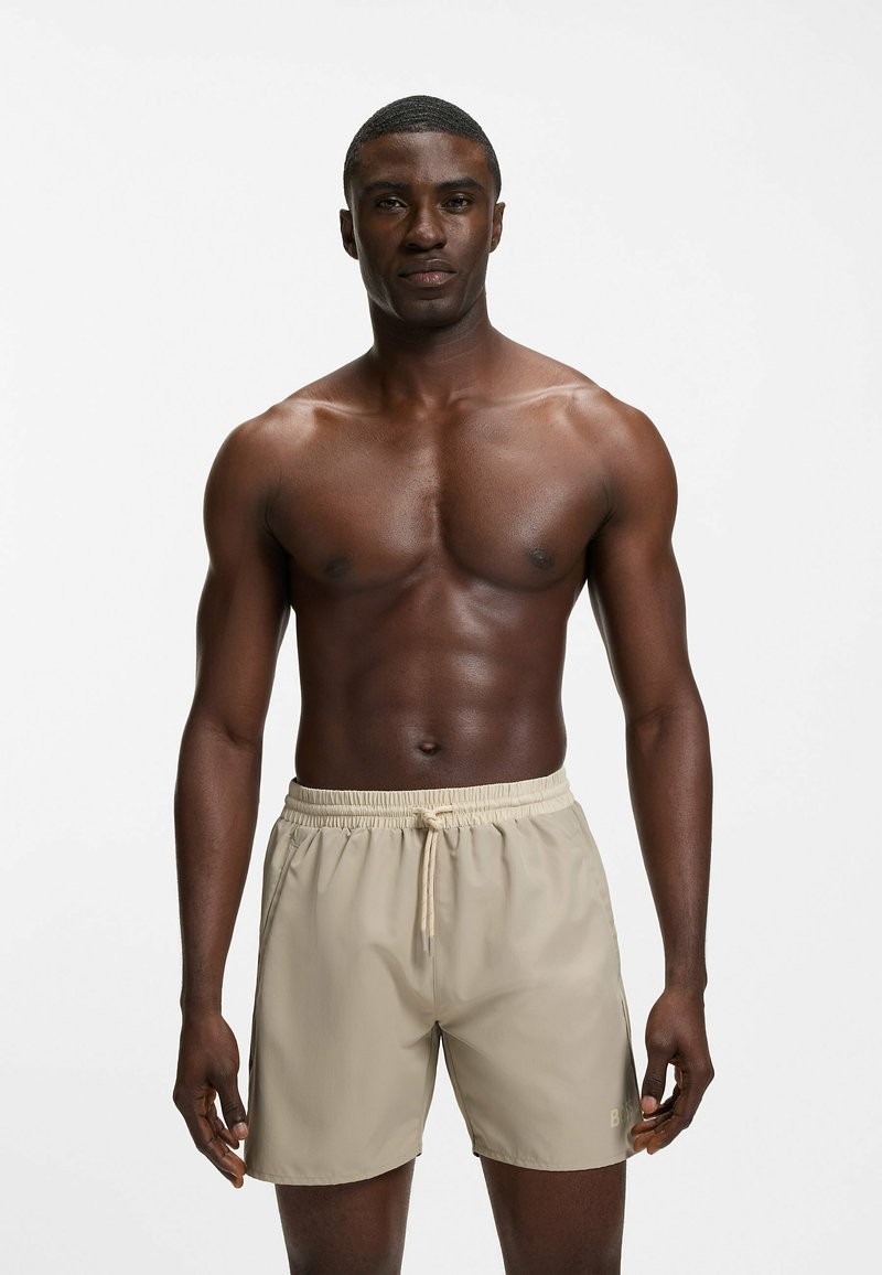 Muscular man standing facing forward wearing beige swim shorts with a drawstring against a plain white background.