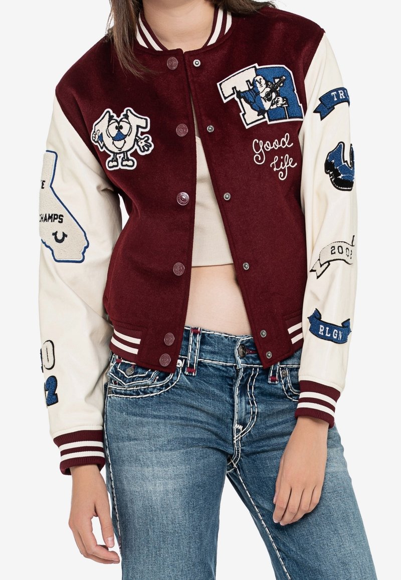 Bordowo-kremowa kurtka varsity z haftowanymi naszywkami, żebrowanymi mankietami i zatrzaskami. Charakteryzuje się sportowym stylem i dekoracyjnymi tekstami.