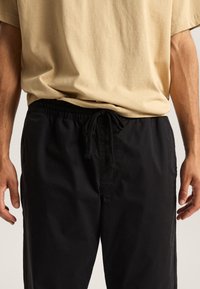 Sorte træningsbukser med snor i taljen, parret med en lys beige t-shirt med korte ærmer, der viser en afslappet pasform og glat tekstur.